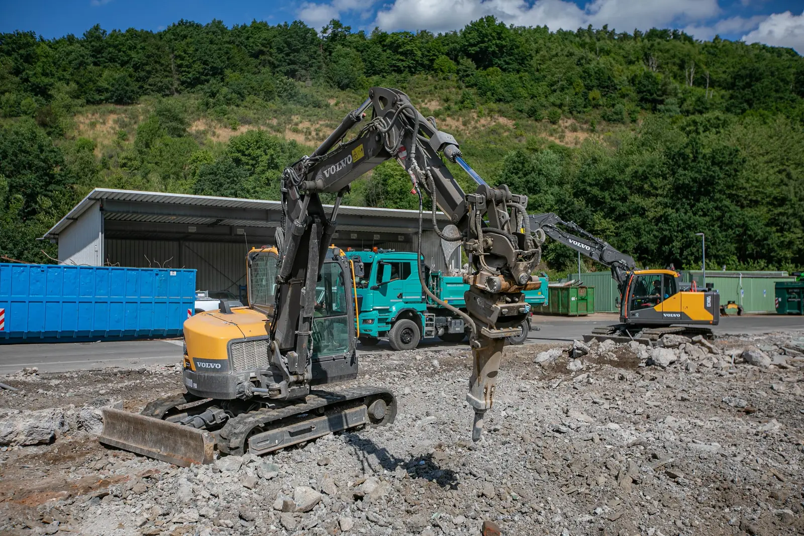 Bagger mit Hydraulikhammer bricht Beton auf einer Industriefläche, dahinter Kipper und ein zweiter Bagger