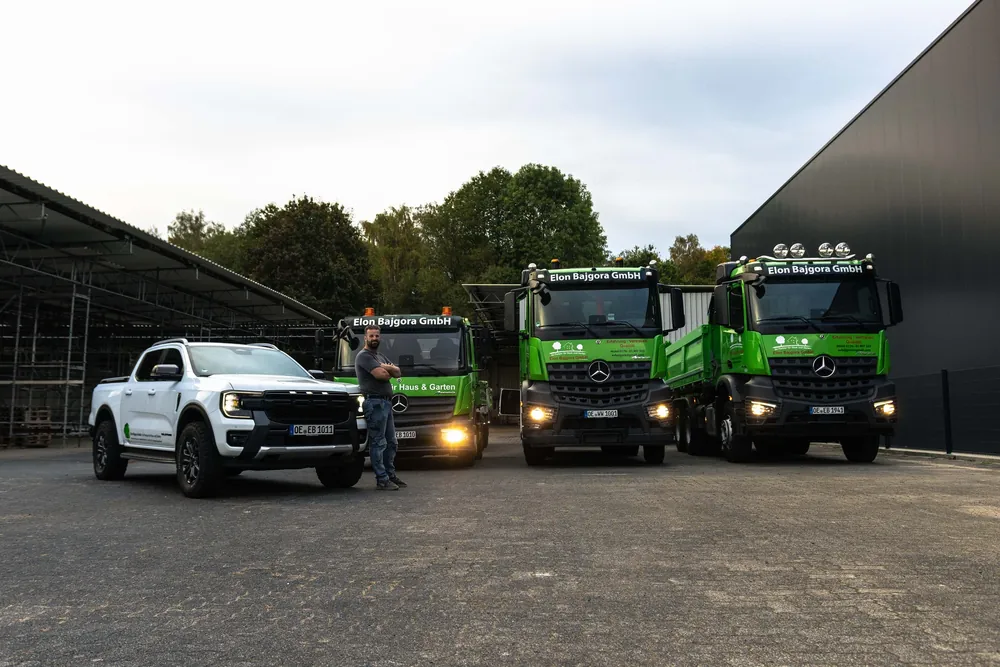 Inhaber Avni Bajgora vor einem weißen Pick-up und den grünen Kippern der Elon Bajgora GmbH